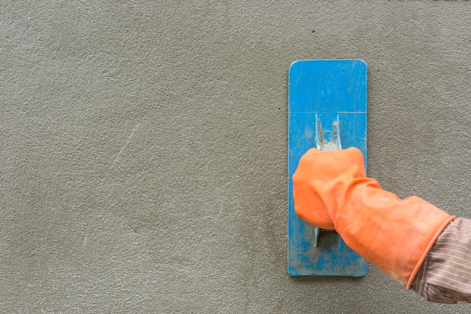 Ouvrier appliquant un enduit de finition sur un mur extérieur avec une taloche bleue, portant des gants de protection orange