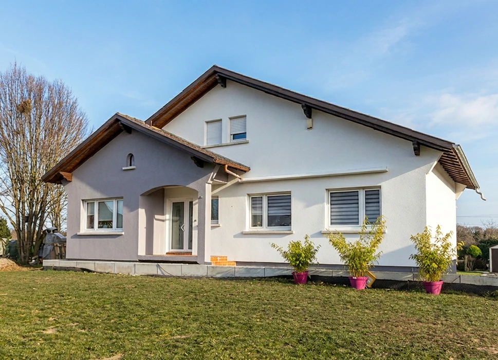 Maison individuelle de plain-pied avec étage partiel, facade en enduit blanc et gris, toiture à deux pans en tuiles sombres avec débords en bois