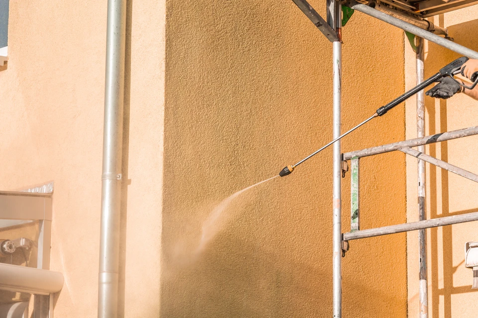 Photographie réaliste en haute résolution d'une façade résidentielle extérieure présentant des signes visibles d'usure et de dégradation : traces de pollution atmosphérique, zones de décoloration irrégulière, légers efflorescences ou taches humides, microfissures discrètes, et altération générale de la finition de crépis ou peinture ancienne. La maison doit être de style classique ou péri-urbain typique de la région Alsacienne. Ciel naturel, éclairage naturel du jour, sans éléments de construction ou outils visibles. L'image doit clairement communiquer le besoin de rénovation de façade.