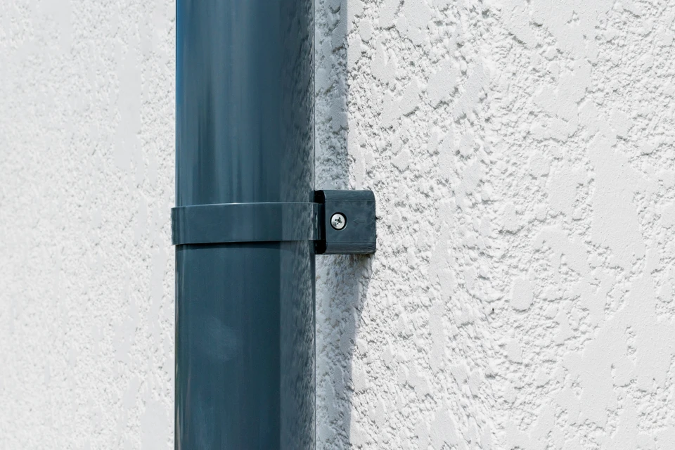 Ouvrier façadier en tenue de travail et équipement de sécurité appliquant un enduit de façade à la truelle sur une paroi extérieure de maison, montrant le geste technique du crépissage professionnel. La scène se déroule en plein jour avec une maison résidentielle française en arrière-plan, capturant le savoir-faire artisanal et la rigueur du travail de façadier.